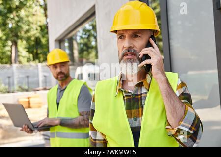 attractive builder with tattoos working on laptop while his colleague talking by phone during work Stock Photo