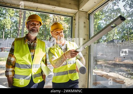 joyful construction workers in safety vests with blueprints and level looking away, builders Stock Photo