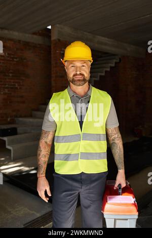 cheerful good looking builder in safety vest and helmet holding toolbox and looking at camera Stock Photo