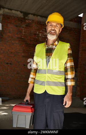 cheerful good looking builder in safety vest and helmet holding toolbox and looking at camera Stock Photo