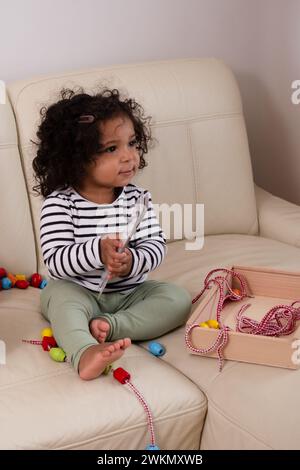 Smiling African American toddler with tousled curls plays with beads on a neutral couch, embodying innocence and engagement. High quality photo Stock Photo