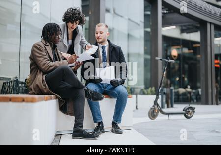 Three young, professional business colleagues collaborating on a project outdoors with paperwork, showcasing teamwork and diversity. Stock Photo