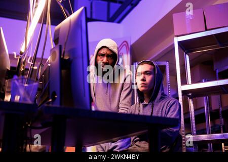 Two concentrated hackers collaborating to crack complex password and break into network system. Young diverse men in hoods working on computer together and coding virus malware Stock Photo