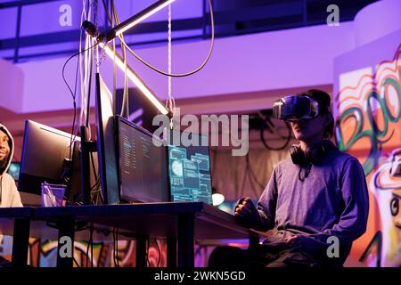 Hacker wearing vr glasses, immersed in simulation while doing illegal activity. Young criminal using virtual reality headset to code malicious software in metaverse in abandoned warehouse Stock Photo