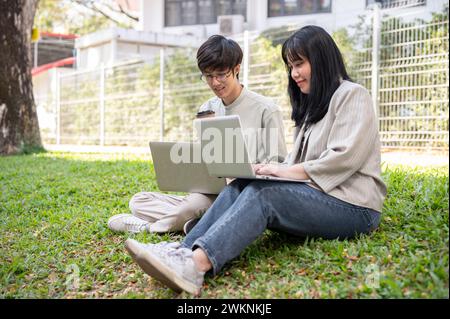 Two young Asian college students are discussing work, sharing their ideas, and using their laptop computer at a campus park. Stock Photo