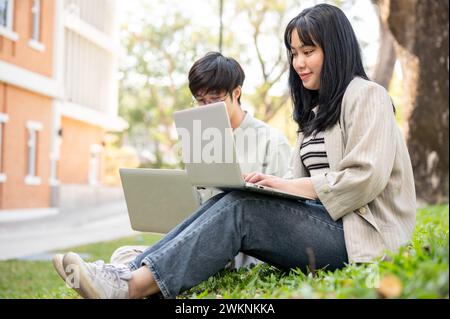 Two young Asian college students are discussing work, sharing their ideas, and using their laptop computer at a campus park. Stock Photo