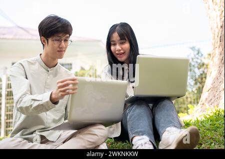 Two Asian university students are collaborating on work, looking at the laptop screen, talking, and sharing their ideas on a project while sitting on Stock Photo