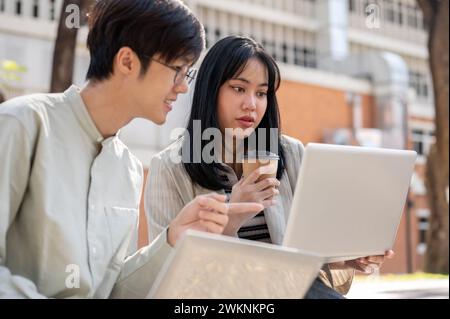 Two smart Asian university students are discussing and collaborating on a project while sitting together on a bench in the university park. university Stock Photo