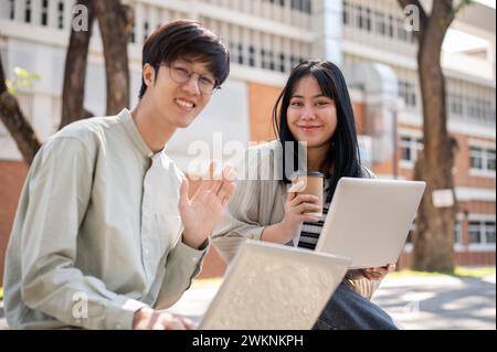 Two smiling Asian university students are sitting on a bench in the university park together with their laptop computer, smiling at the camera. Stock Photo