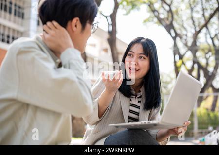Two smart Asian university students are discussing and collaborating on a project while sitting together on a bench in the university park. university Stock Photo