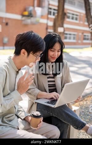 Two smart Asian university students are discussing and collaborating on a project while sitting together on a bench in the university park. university Stock Photo
