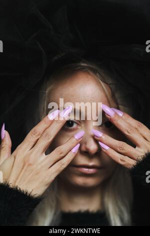 A striking image of a young blonde caucasian woman with purple nails covering her face with both hands, wear a vintage black lace dress and hat Stock Photo