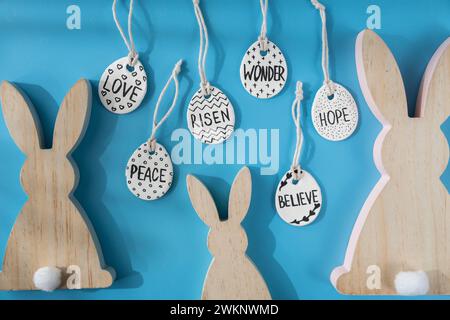 Group of wooden bunny ears and clay Easter eggs with words RISEN HOPE BELIEVE PEACE LOVE WONDER on blue background. Children air dry clay activity handicraft idea. Preparing for Easter holiday decorating. Modern organic design minimalistic plastic free sustainable decor  Stock Photo
