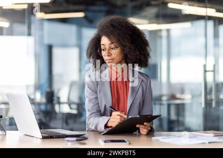 Focused female in round eyeglasses looking at pc screen while filling in document on writing board in office interior. Confident entrepreneur copying information from email to printed papers. Stock Photo