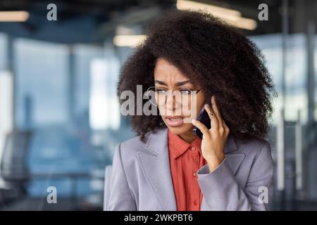 Worried african american lady with bushy hairstyle holding smartphone by ear and listening bad news. Disturbed office worker having stressful distant conversation by mobile gadget during daytime. Stock Photo