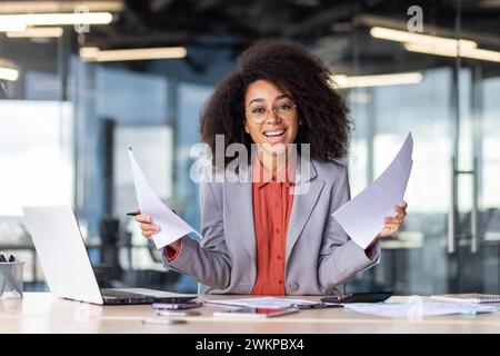 Positive african american employee doing paperwork while sitting by portable computer in modern workspace. Smiling lady holding documents in hands while looking at camera during busy day indoor. Stock Photo