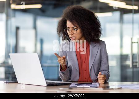 Angry head of department looking at laptop screen and pointing with finger at webcam while sitting by desk in cabinet. Disappointed boss scolding subordinate colleague for poor work efficiency. Stock Photo
