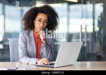 Thoughtful latino person resting head on chin while working by portable computer next to tablet and notebook. Pensive female boss making choice which employee deserving promotion to leading position. Stock Photo