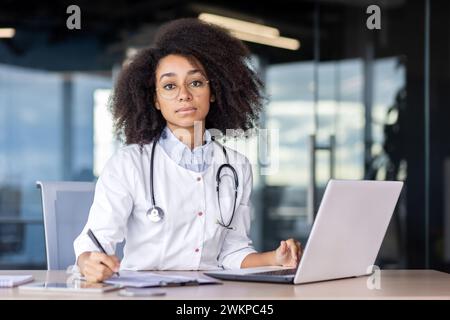 Attentive female doctor in white uniform writing with pen on clipboard while looking at camera. Focused therapist sitting by desktop and making prescription for medication at specialised pharmacy. Stock Photo