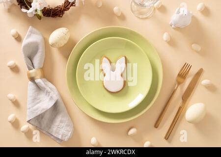Easter table setting with white and green plate, bunny cookies, eggs on beige background. View from above. Happy Easter. Stock Photo
