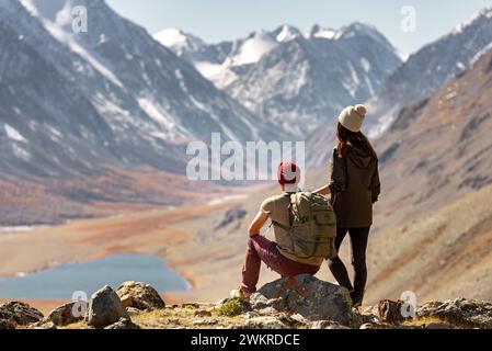 Couple of tourists with backpack are relaxing at observation point high in mountains and enjoys the view at lake and glaciers Stock Photo