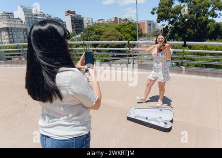 Unrecognizable woman is filming with her phone busker female violinist outdoors while playing violin and making street music, technology concept, copy Stock Photo