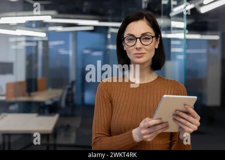 A professional woman confidently holds a tablet in a modern office environment, displaying poise and expertise. Stock Photo