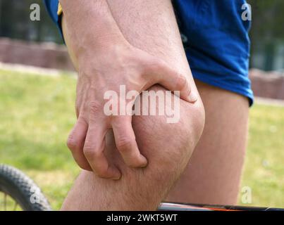 Young man in sportswear on bike in urban park touching his painful knee Stock Photo