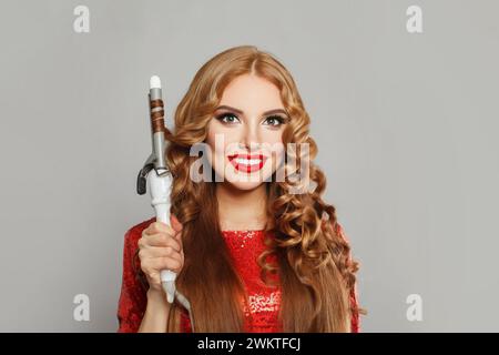 Happy young glamorous woman with long straight and curly hair holding curling iron on white background. Hair care, hair styling and beauty concept Stock Photo