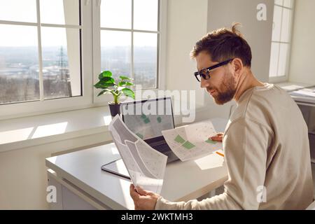 Portrait of serious, focused, attractive architect in process of work, sitting at written desk at his workplace in office. Stock Photo