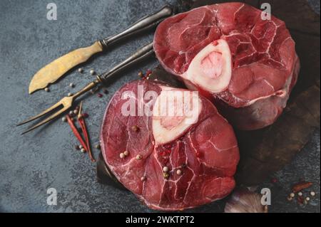 raw Ossobuco. Veal shanks Ossobuco with pepper, lemon and garlic for gremolata, typical ingredients of Italian Lombard cuisine, top view on dark backg Stock Photo
