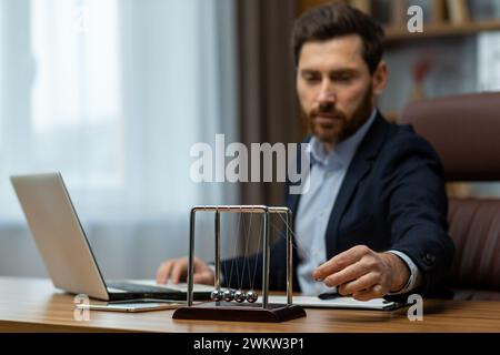 A serious and successful young man sitting in the office at a desk in a suit uses ,Newton's Cradle by launching balls with his hand. Stock Photo