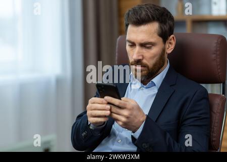 Concentrated and serious young businessman sitting in a chair in the office at the table and using a mobile phone, typing messages, texting, reading n Stock Photo