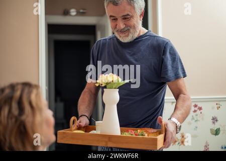 In this close-up shot, we witness a heartwarming moment between a retired couple. The husband stands by the bedside, tenderly holding a tray laden wit Stock Photo