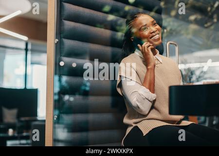 Successful young black businesswoman sits in a coworking space, speaking on a smartphone with a happy smile. She embodies professionalism and remote w Stock Photo