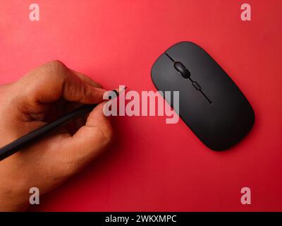 Someone hand holding pencil with black wireless mouse on a red background. Stock Photo