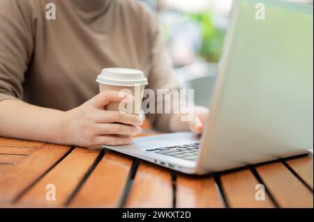 A close-up shot of a woman holding a takeaway coffee cup, drinking coffee while working on her laptop computer at a cafe in the city. working remotely Stock Photo