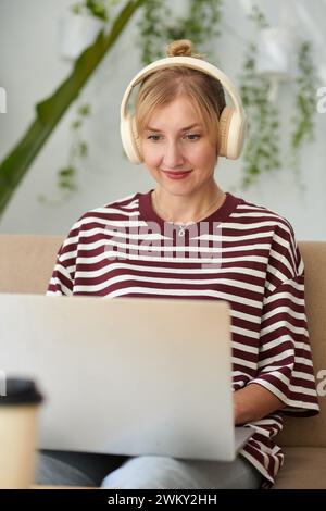 Portrait of smiling woman listening to music in headphones when coding on laptop at home Stock Photo