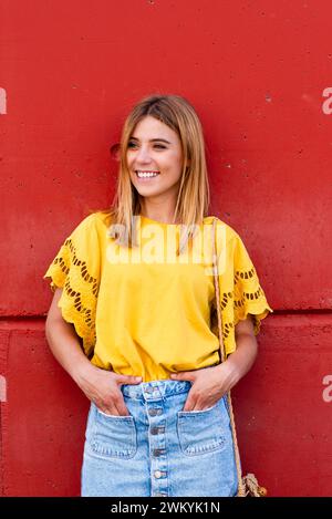 A young woman stands confidently against a red wall, her yellow blouse Stock Photo