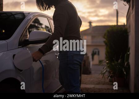 Man plugs a charge cable into the electric car in the house area at evening. Concept of green energy and sustainability. Stock Photo
