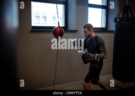 Concentrated young adult bearded boxer honing skills on double-end punching bag, working out focus and accuracy in dimly boxing gym Stock Photo