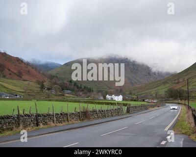 Backed by hills under a cloudy sky a white open topped sports car drives along the A592 nr Ullswater in the National Park. Stock Photo