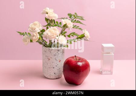 Bouquet of white carnations in a vase, a red apple and a bottle of perfume on the pink background. Stock Photo