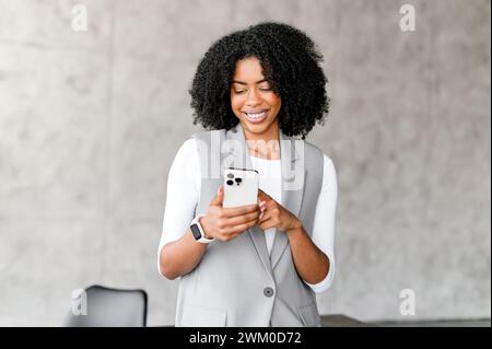 An engaging African-American businesswoman interacts with her smartphone, her expression reflecting the ease and efficiency of modern business communications within a contemporary office setting. Stock Photo