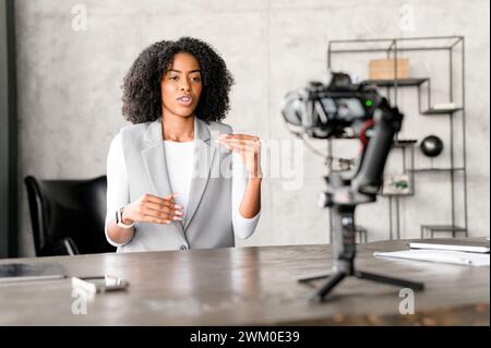 An African-American businesswoman engages confidently with a camera, gesturing as if explaining a complex concept during a video blog or webinar session in a modern office environment. Stock Photo