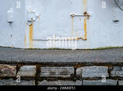 A close-up view of a weathered exterior wall featuring rust stains seeping from metal pipes, peeling white paint, and a textured interface Stock Photo