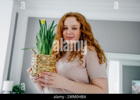 Redhead woman holding daffodil plant in wicker basket at home Stock Photo