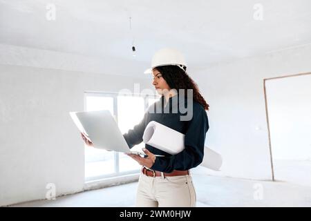 Young architect examining blueprints and standing with laptop at site Stock Photo