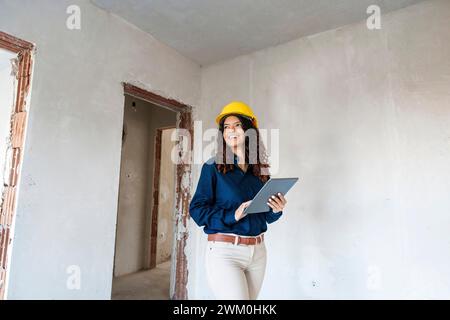 Smiling architect wearing yellow hardhat and standing with tablet PC at site Stock Photo
