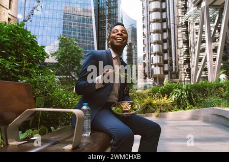 Happy businessman sitting with bowl of salad on bench at office park Stock Photo